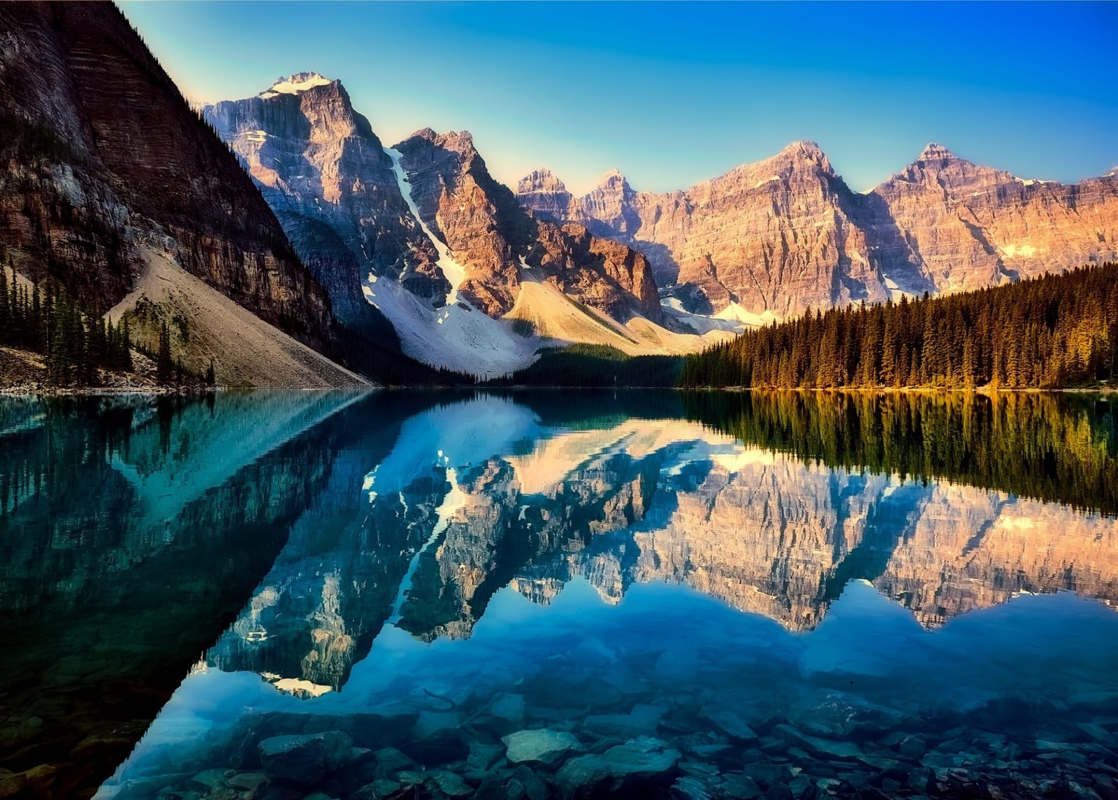 A glacial lake at sunrise, mirror still, with snow-streaked mountain peaks lit warm above a dark band of pines, and turquoise water running clear over pale stones.